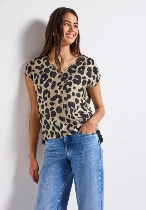 Frau mit Leopardenmuster-Shirt und blauer Jeans, steht lächelnd an einer weißen Wand.